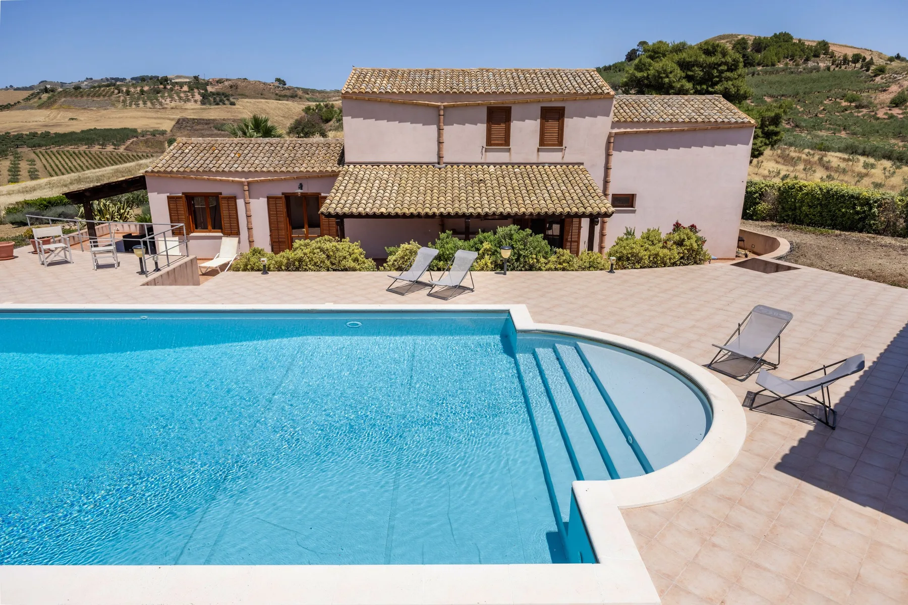 A swimming pool and surrounding patio sit in front of a hillside house.
