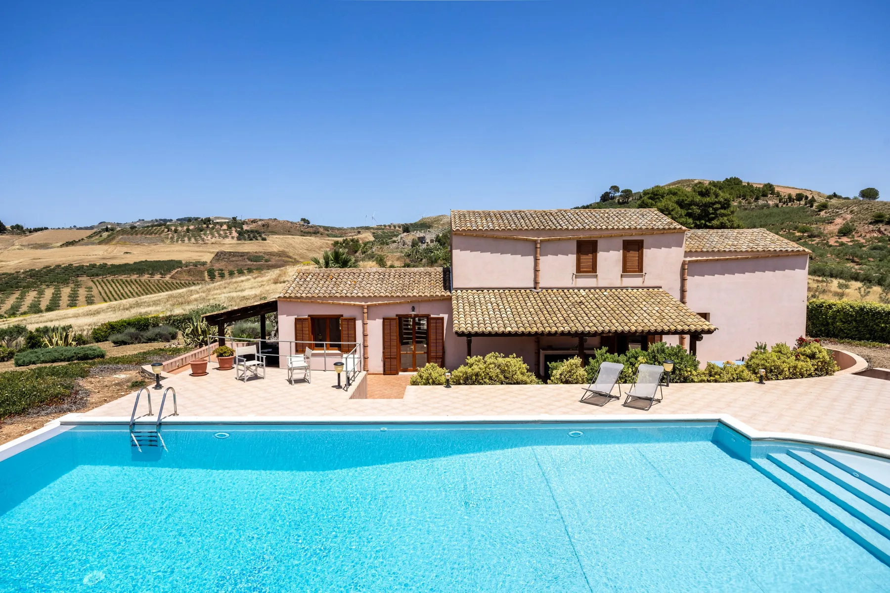 A countryside house overlooks a swimming pool, with lounge seating on the patio.