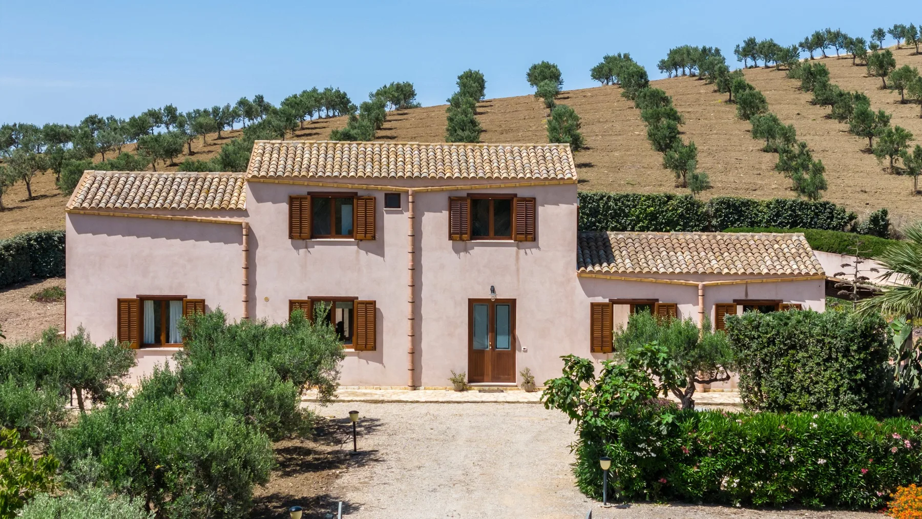 A pale stucco house with tiled roofs sits among shrubs on a dry hillside.