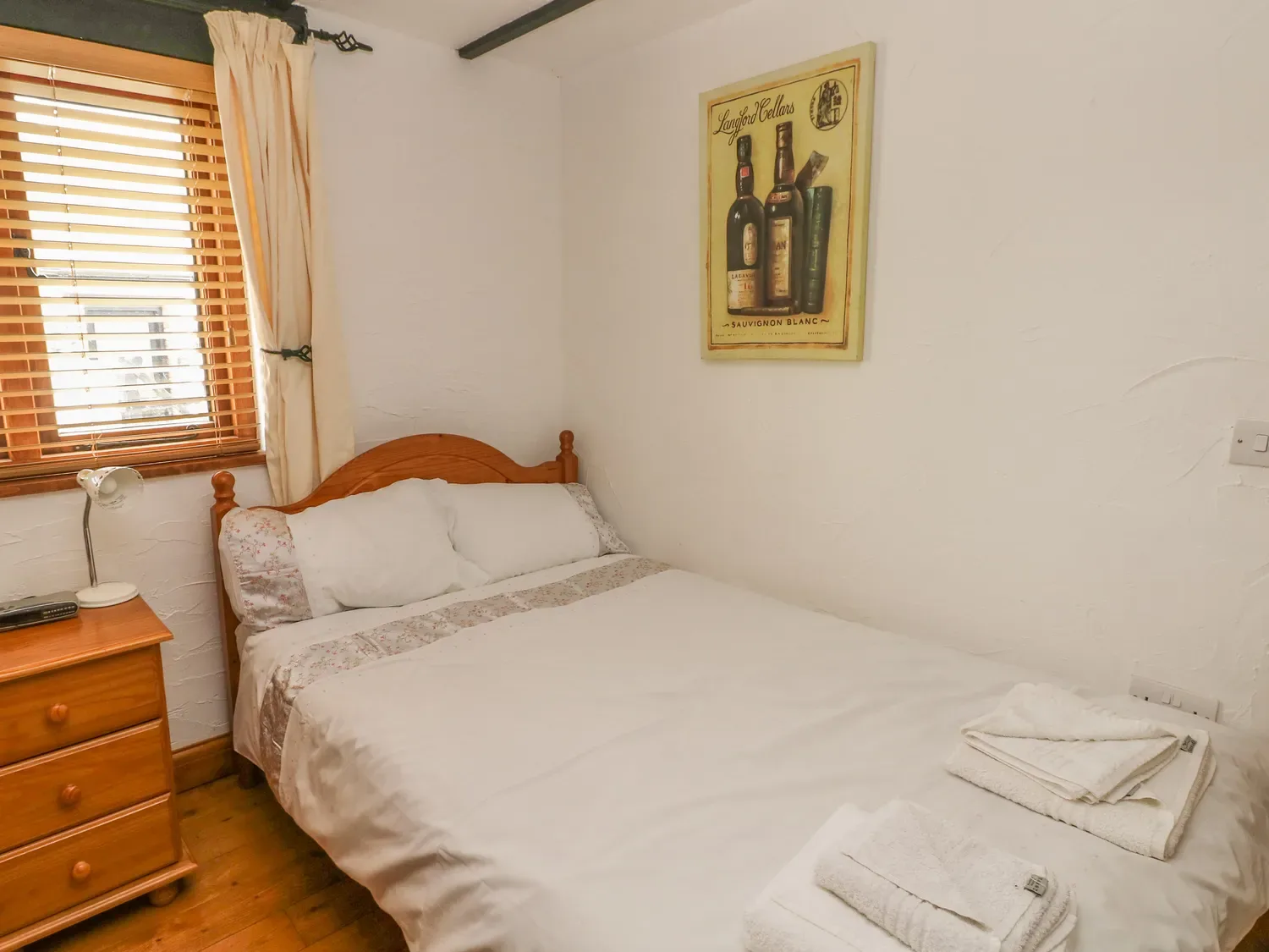 Un dormitorio con una cama de matrimonio, una mesita de noche y estores en la ventana en Cowslip Cottage, en Rosemarket.