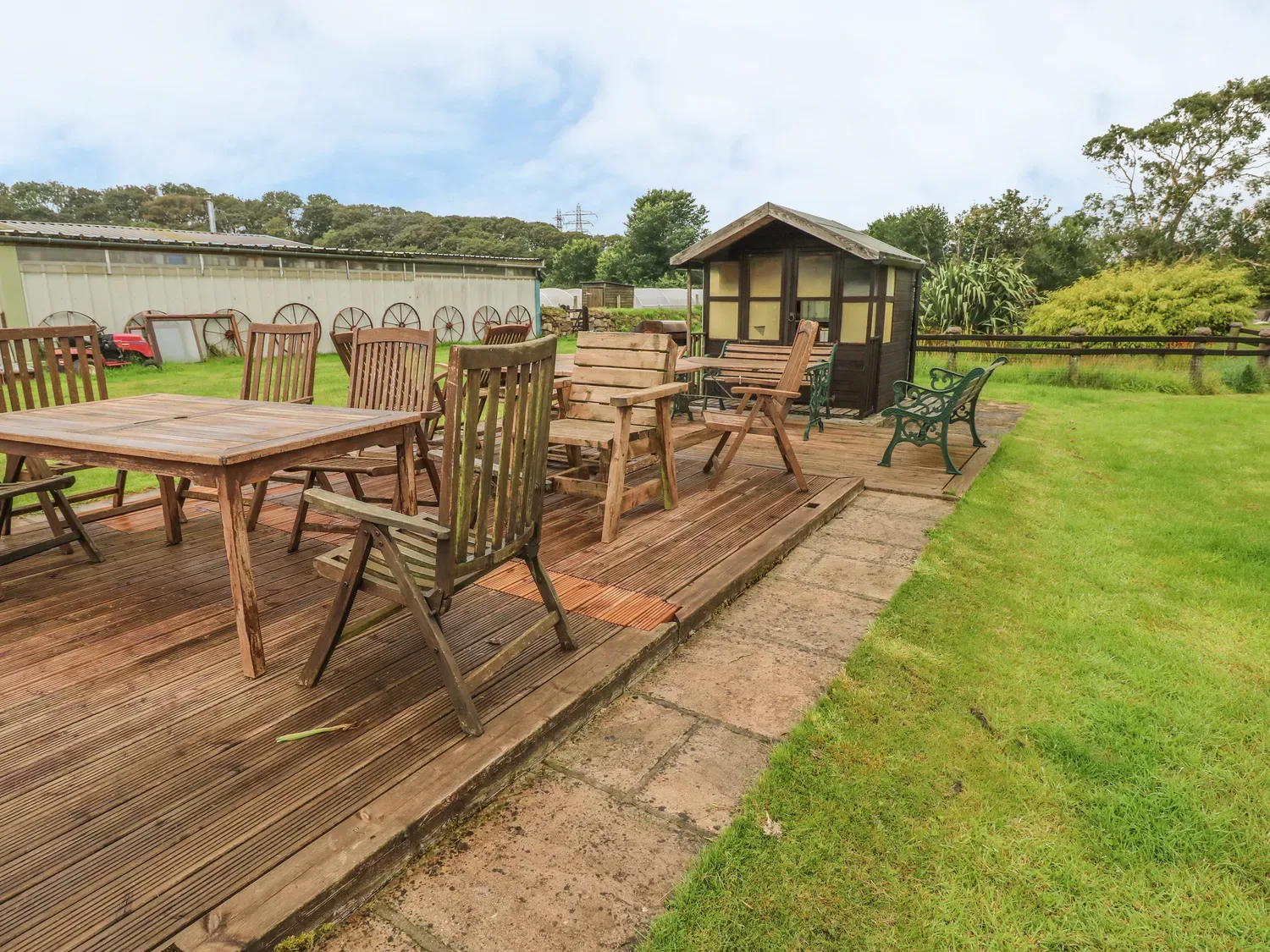 Una terraza exterior de madera con varias sillas y mesas de madera cerca de un pequeño cobertizo y césped verde en Cowslip Cottage, en Rosemarket.
