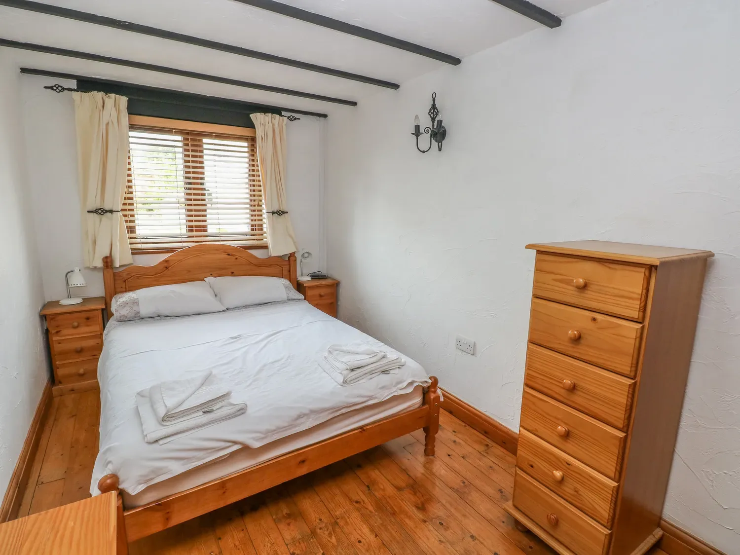 Un dormitorio con una cama de madera, ropa de cama blanca, una cómoda de madera y una ventana con cortinas en Cowslip Cottage, en Rosemarket.