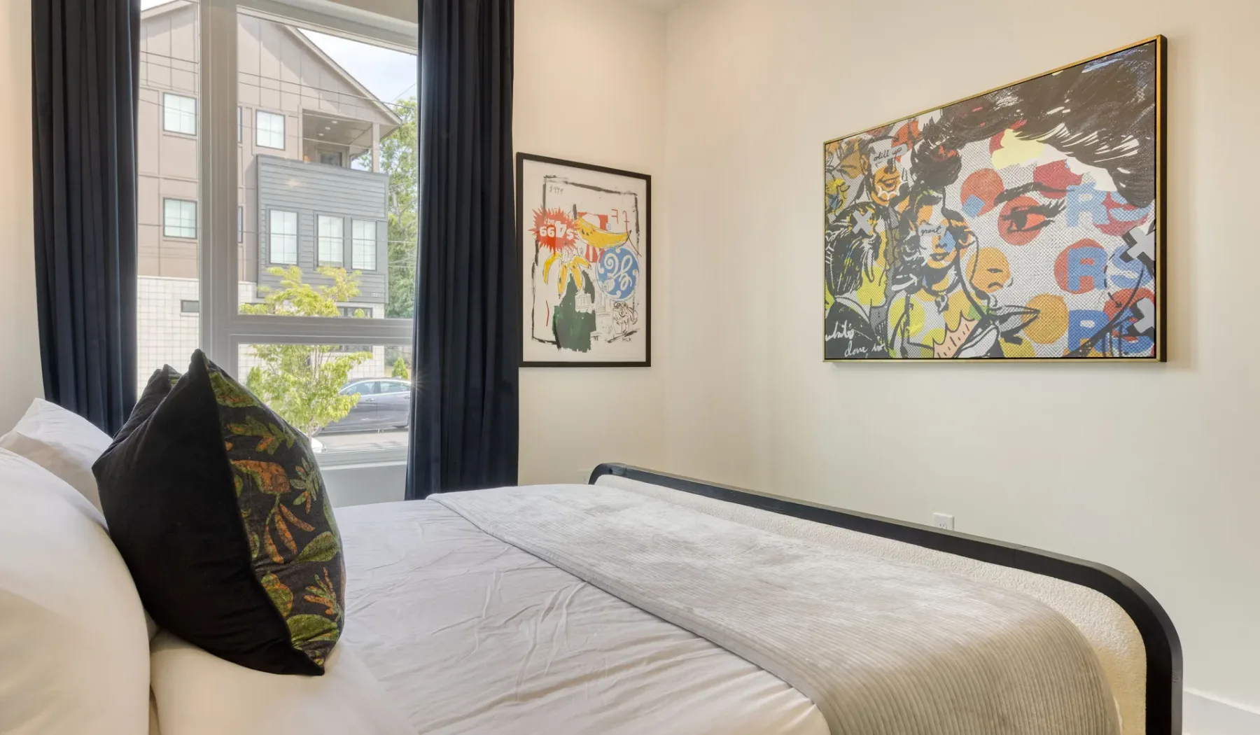A bedroom with a bed, dark curtains, and colorful framed artwork.