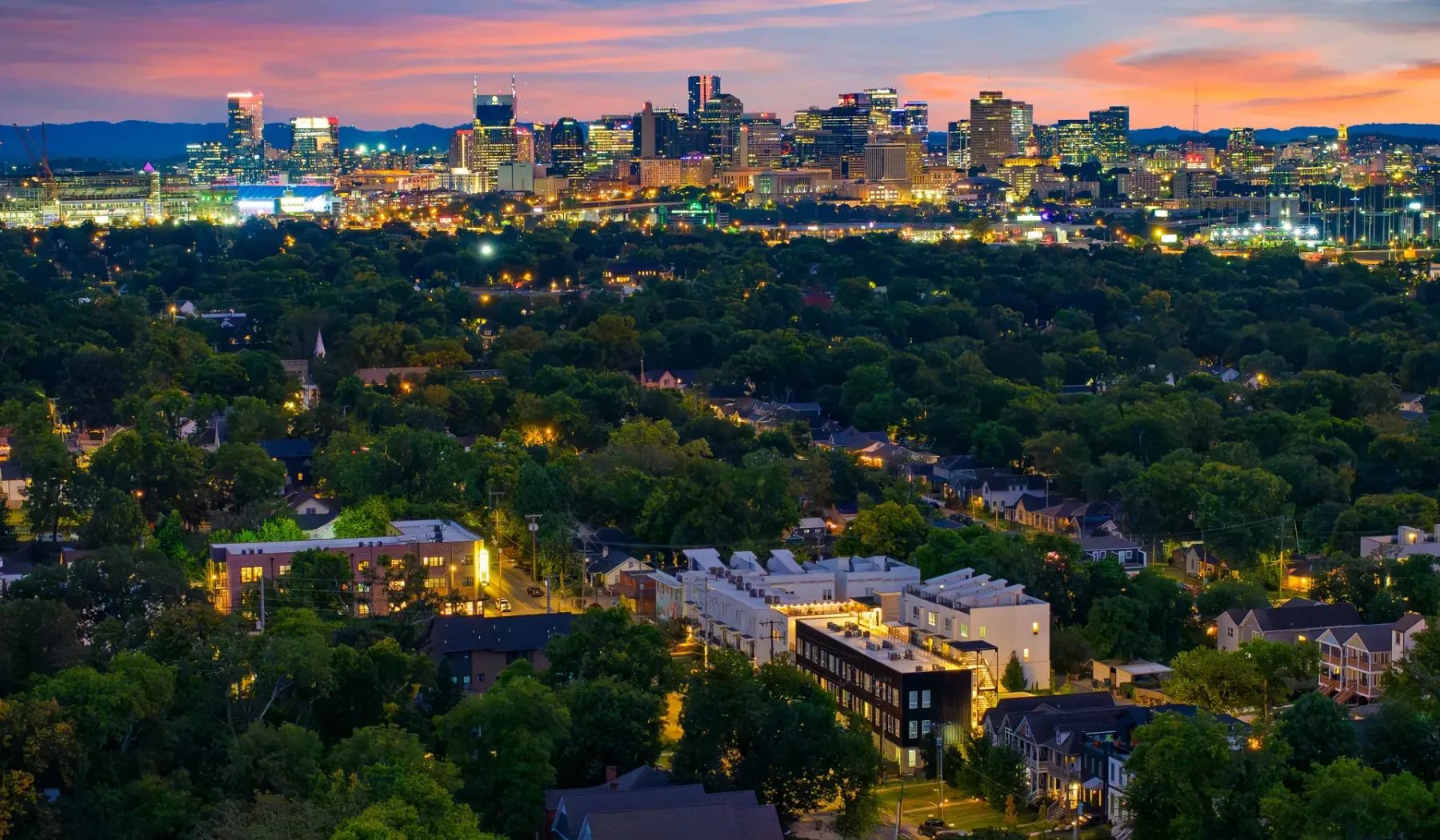 A city skyline glows at dusk beyond a broad stretch of trees.