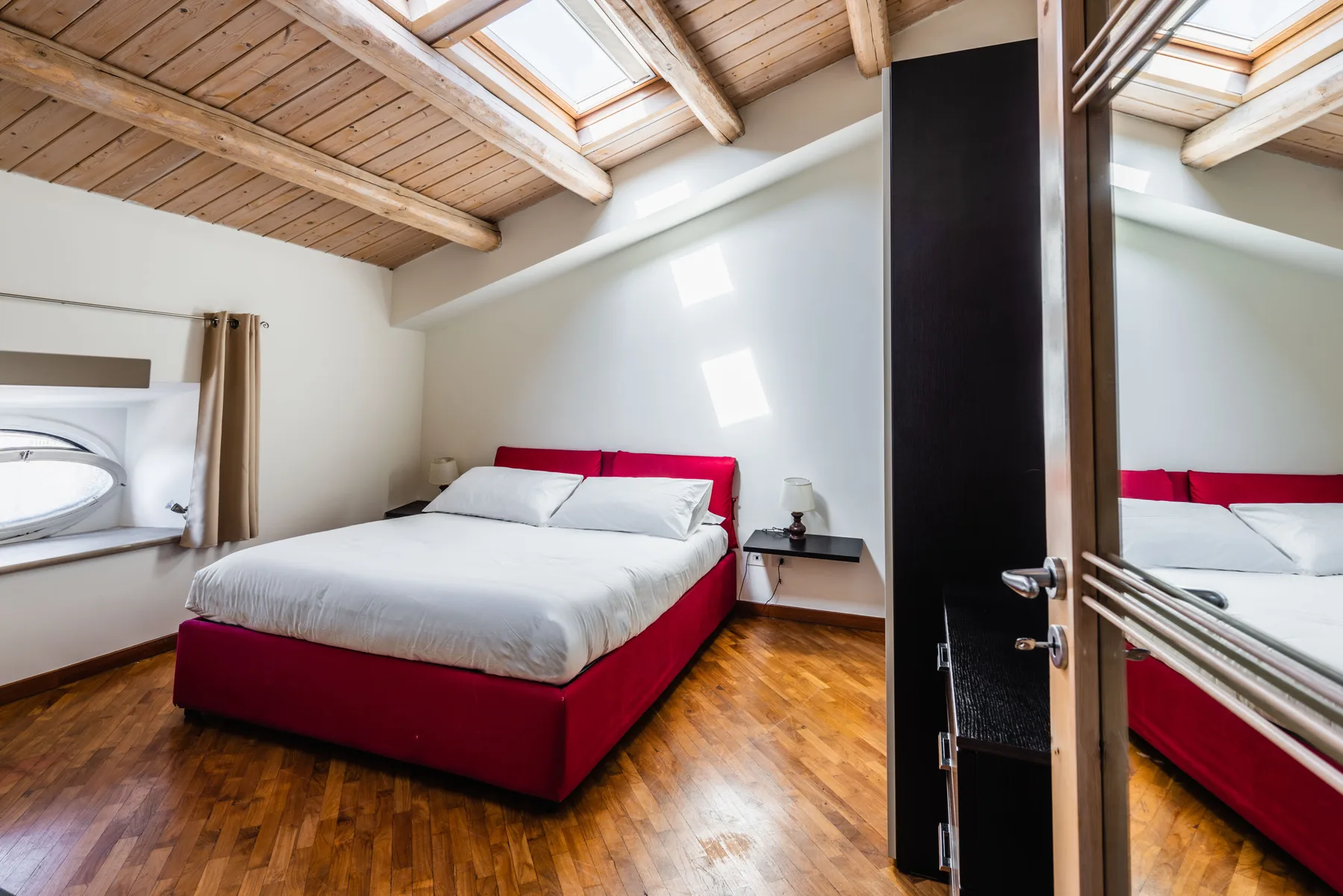 Un dormitorio con una cama roja bajo un techo inclinado de madera y tragaluces.