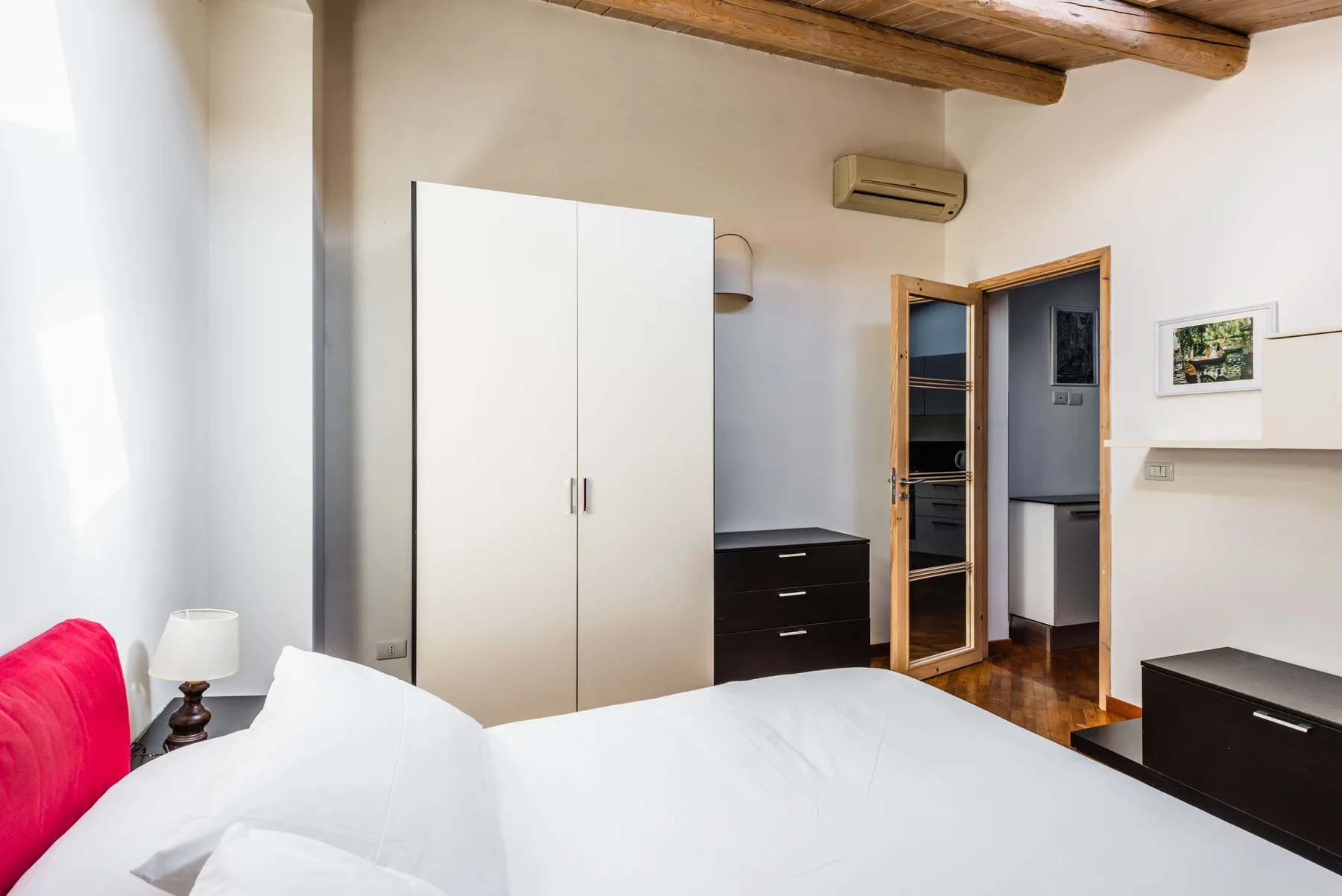 Un dormitorio luminoso con una cama blanca, un armario y vigas de madera oscuras en el techo.