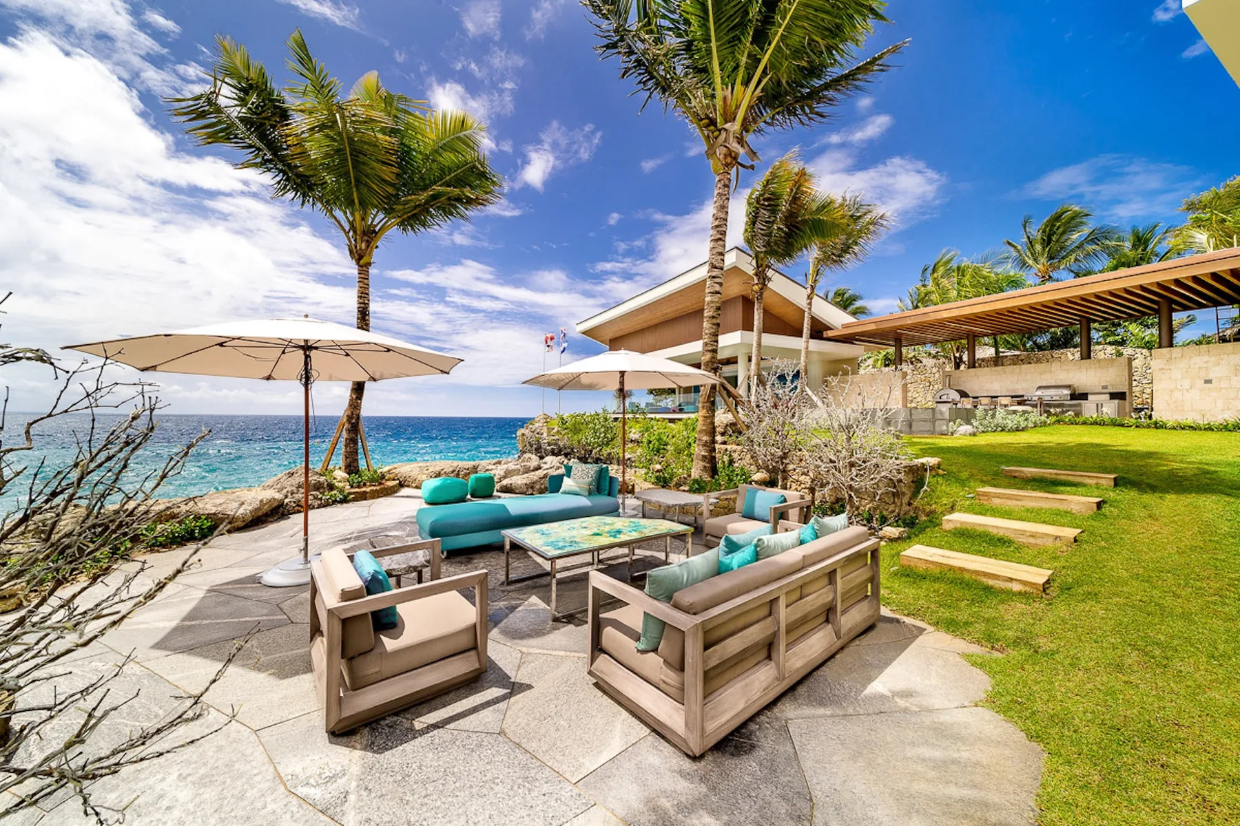 Outdoor seating on a paved terrace beside a lawn, with palm trees and the ocean beyond.