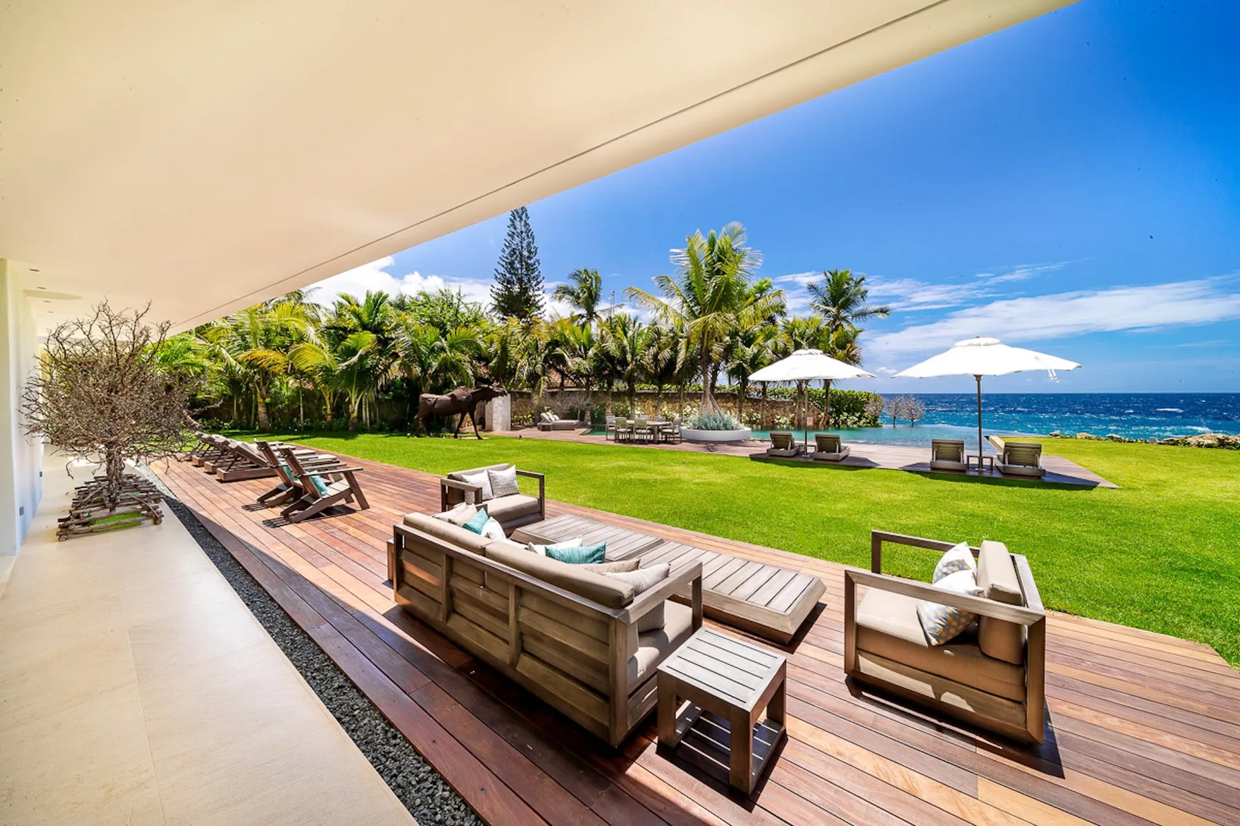A seaside patio with outdoor seating overlooks a lawn, palm trees, and the ocean.