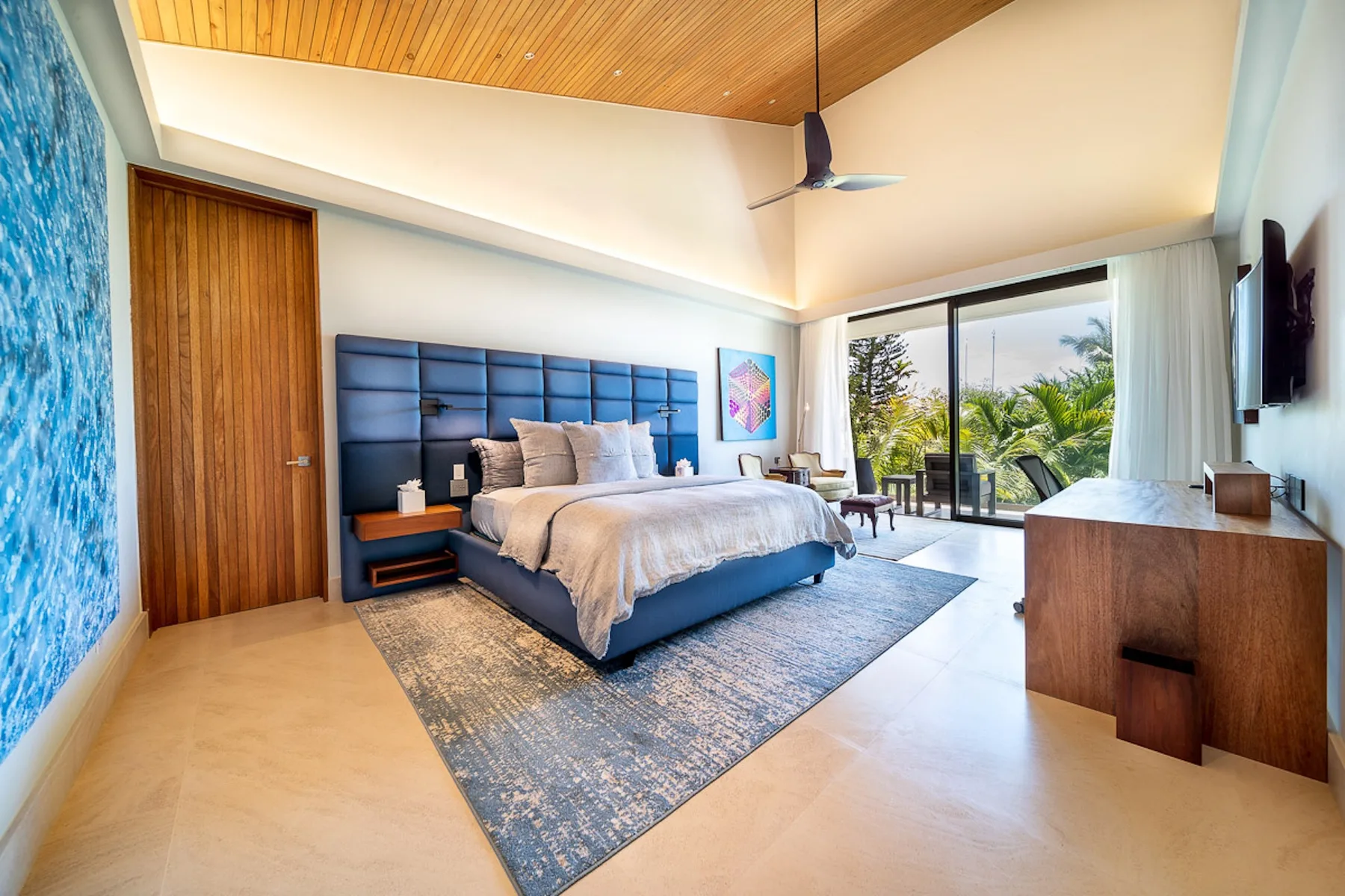 A bedroom with a bed, blue headboard, and sliding glass doors opening to greenery.