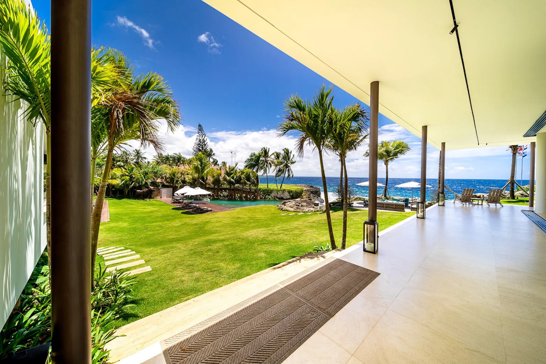 A covered patio overlooks a green lawn, palm trees, and the ocean.