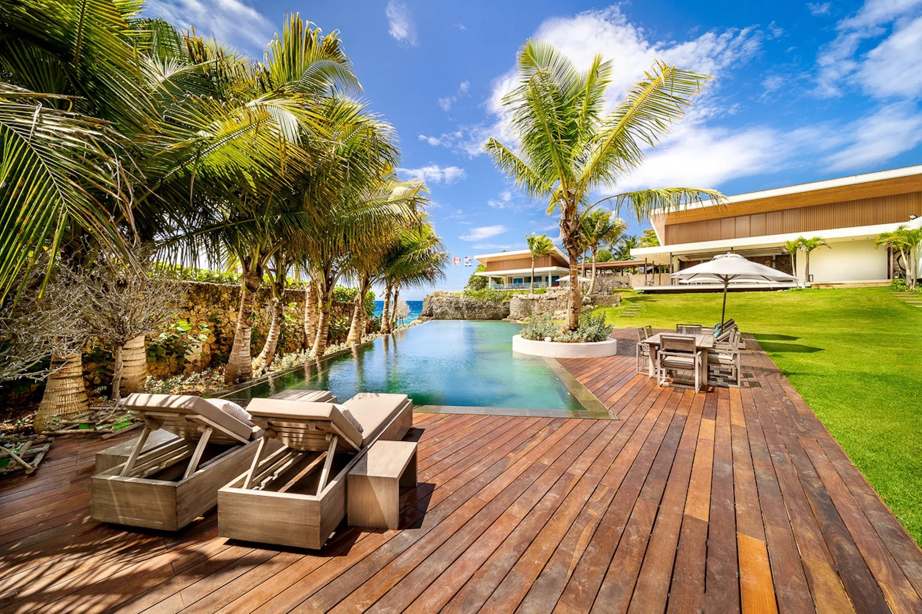 A tropical outdoor pool beside a wooden deck, lounge seating, and palm trees.