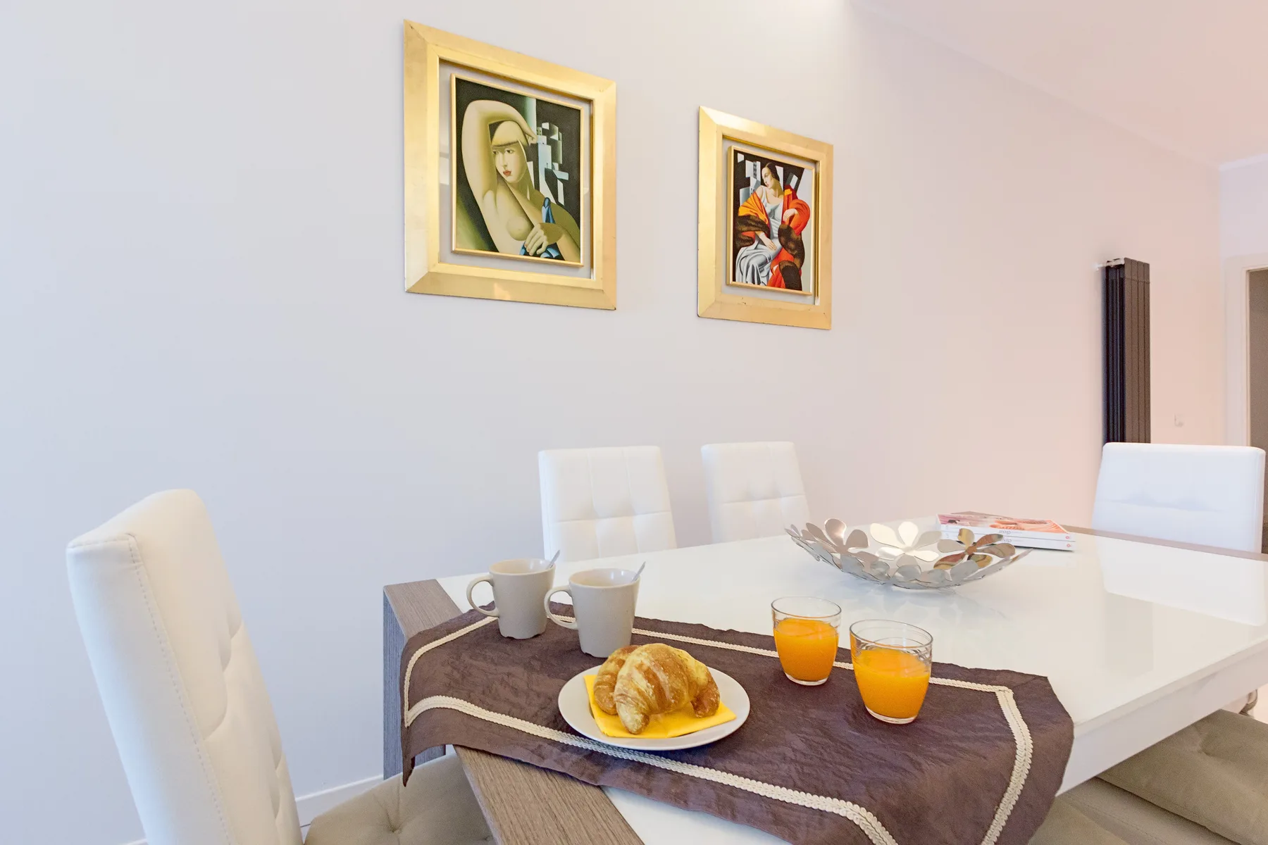 Un comedor con una mesa preparada con bollería y bebidas bajo cuadros enmarcados.