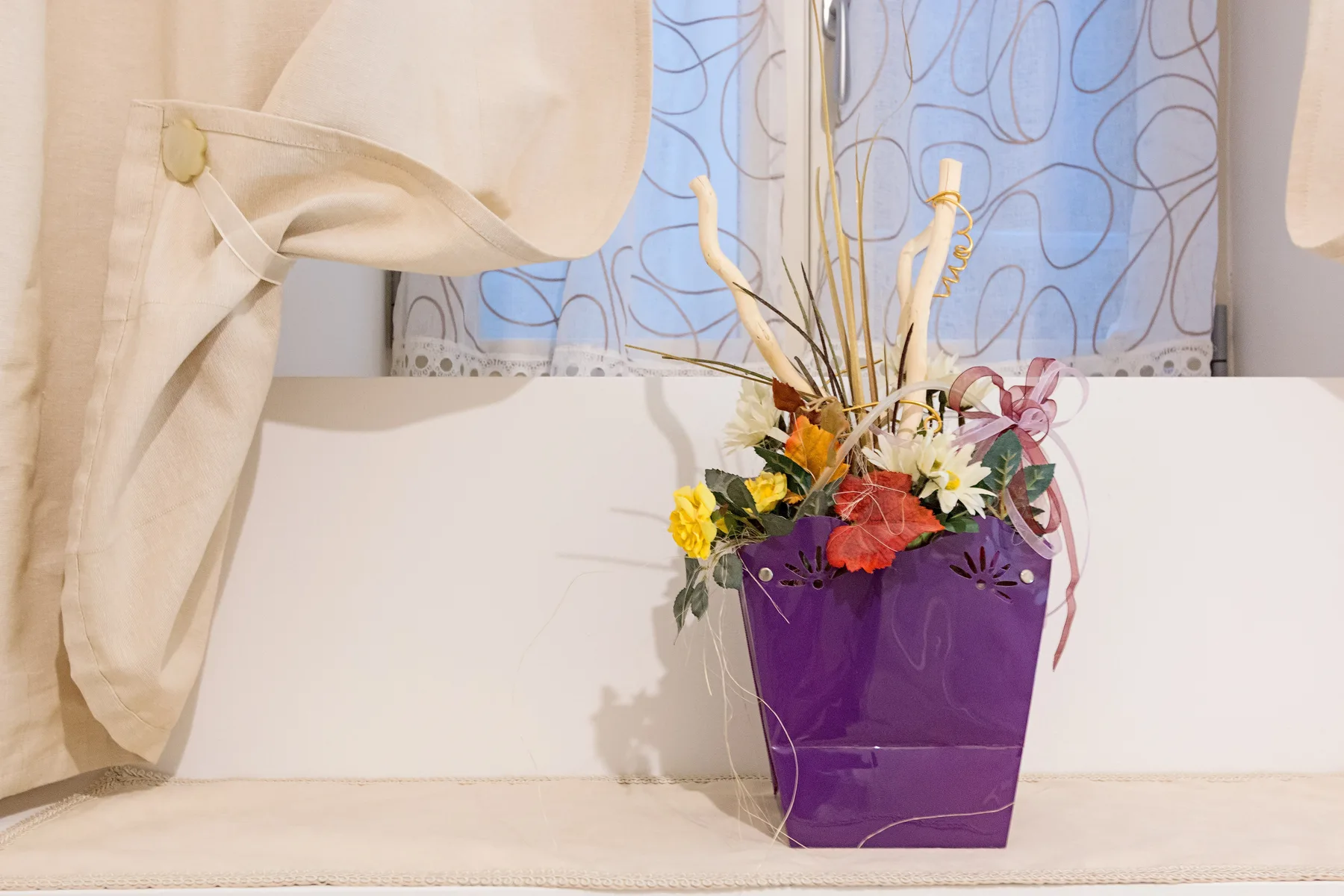 Un colorido arreglo de flores está en un recipiente morado sobre el alféizar de una ventana.