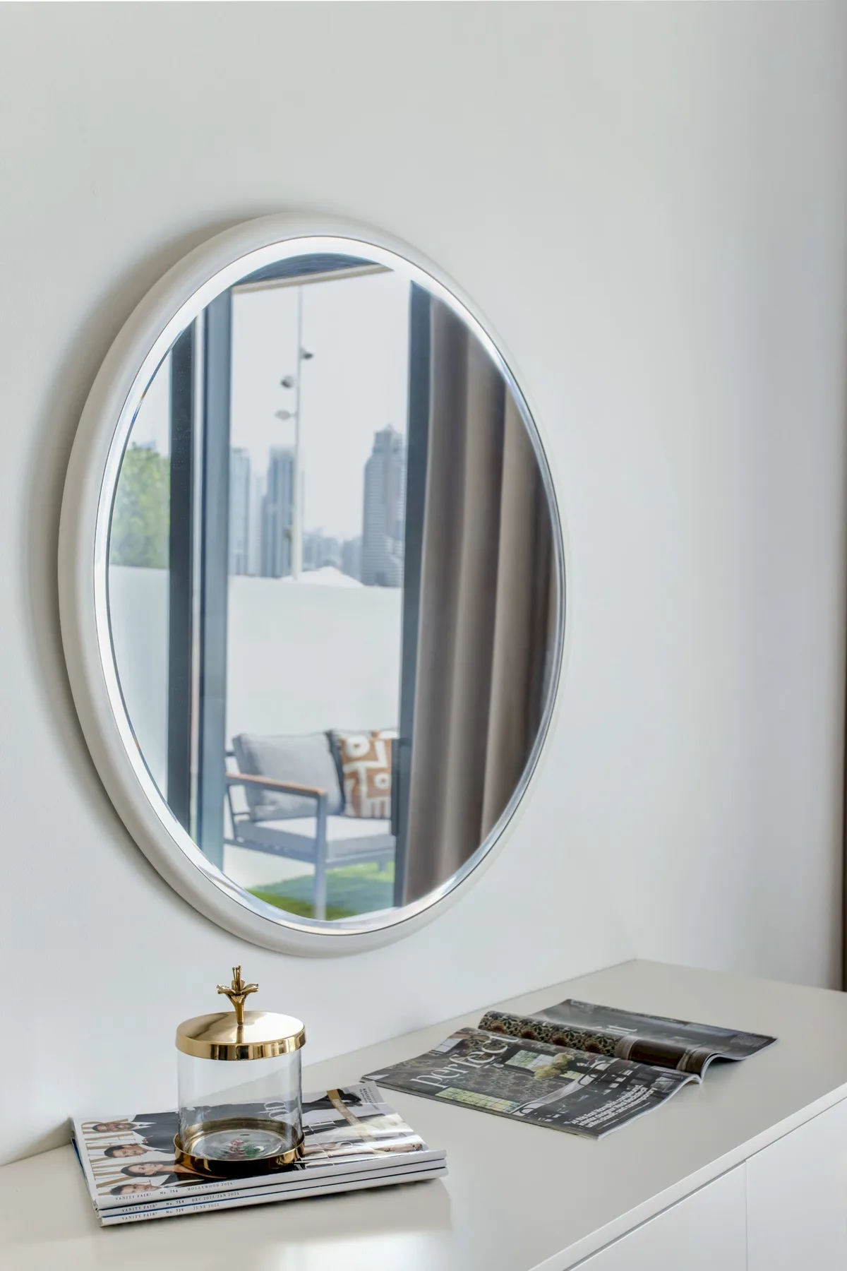 Una cómoda con un espejo ovalado y revistas refleja una habitación luminosa.