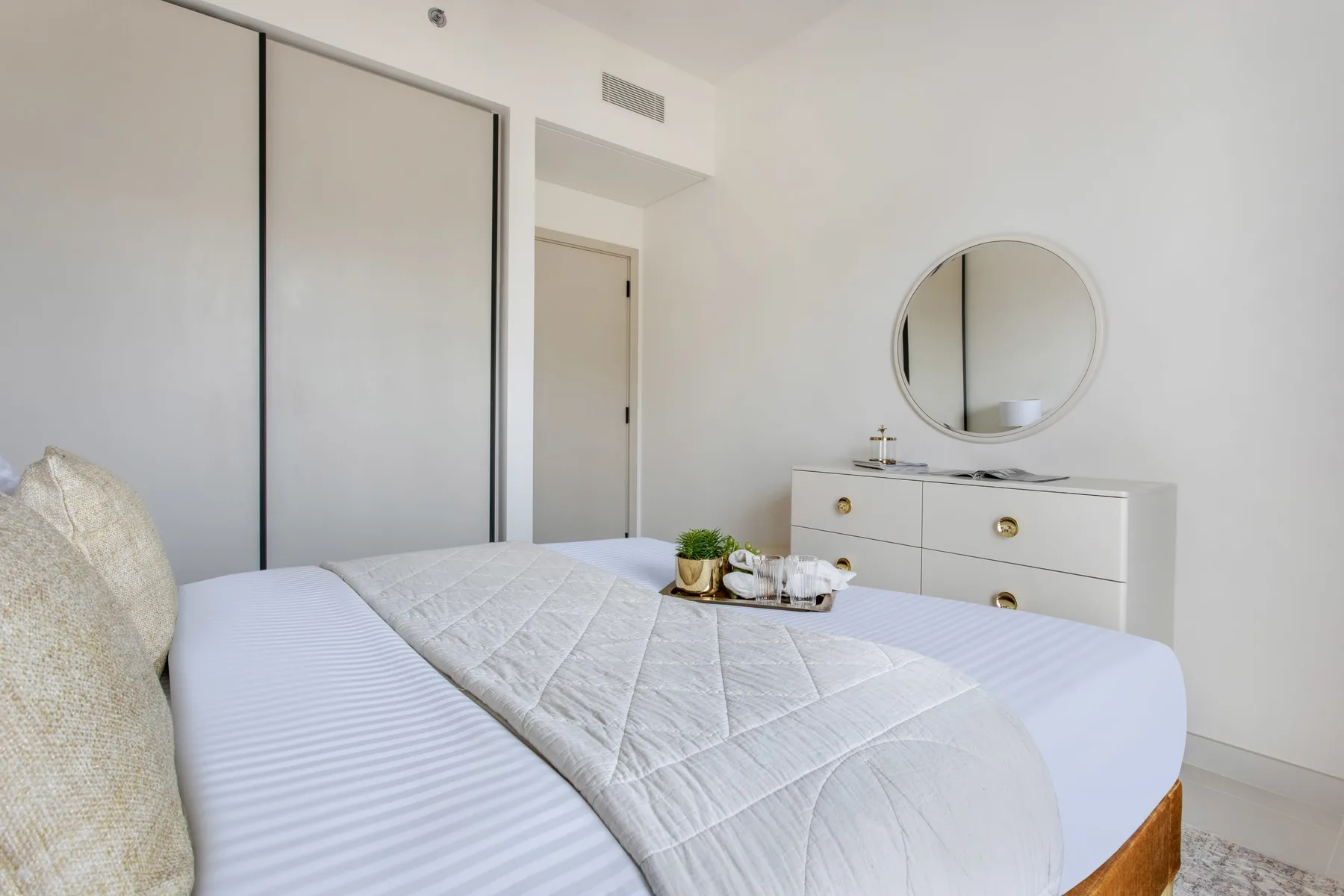 Un dormitorio luminoso con una cama bien tendida y una cómoda con un espejo redondo.