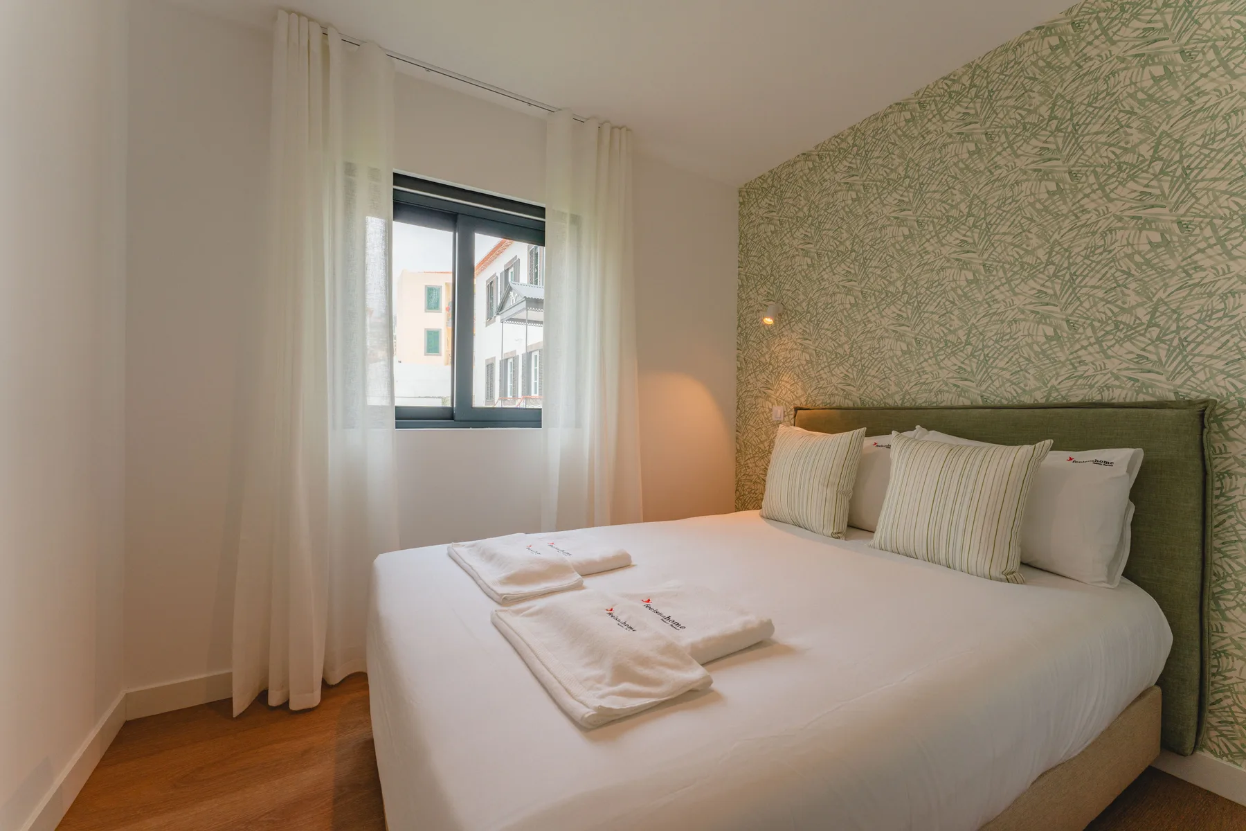 Un dormitorio con una cama bien tendida junto a una ventana con cortinas transparentes.