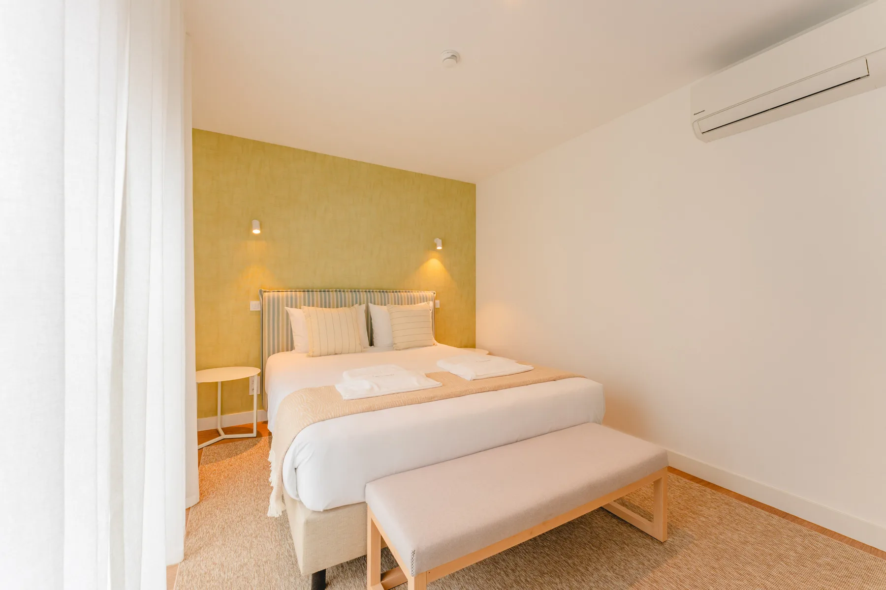 Un dormitorio con una cama bien arreglada, un banco y un aire acondicionado en la pared.