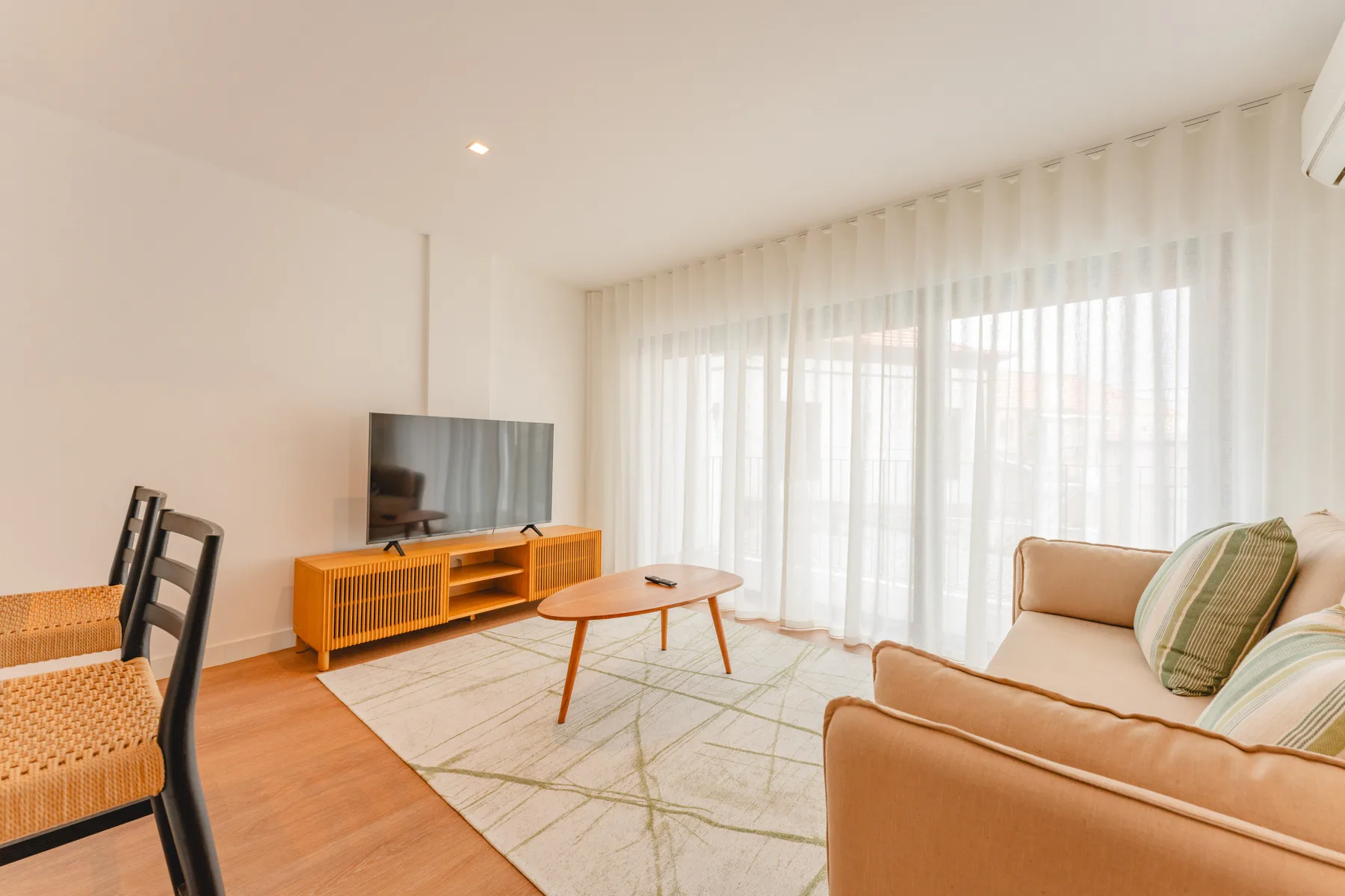 Una sala luminosa tiene asientos, un televisor y cortinas translúcidas frente a las ventanas.