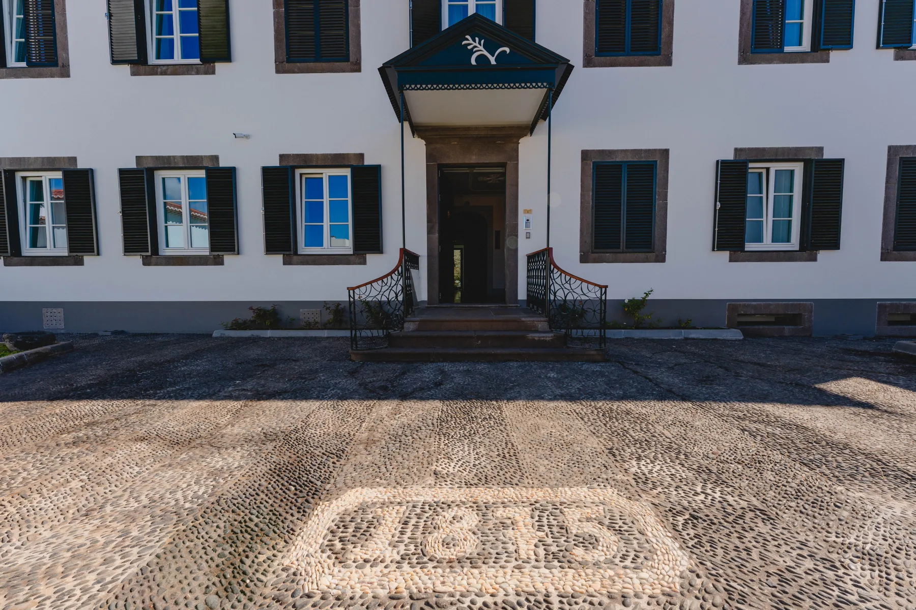 La entrada de un edificio blanco con contraventanas oscuras da a un patio empedrado.