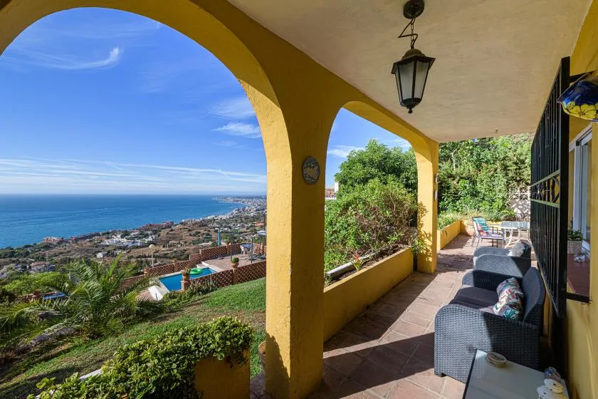 A covered patio with seating overlooks the sea and coastal hillside.