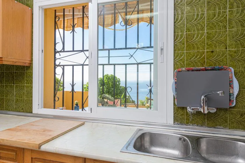 A kitchen sink sits beneath a window overlooking greenery and the water.