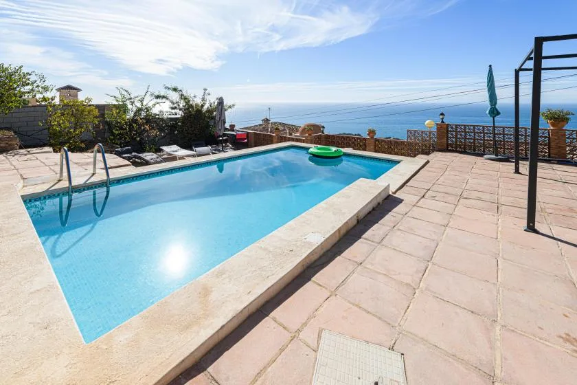 An outdoor swimming pool sits beside a paved terrace overlooking the sea.