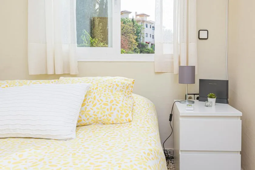 A bright bedroom with a bed beside a white bedside table and window.