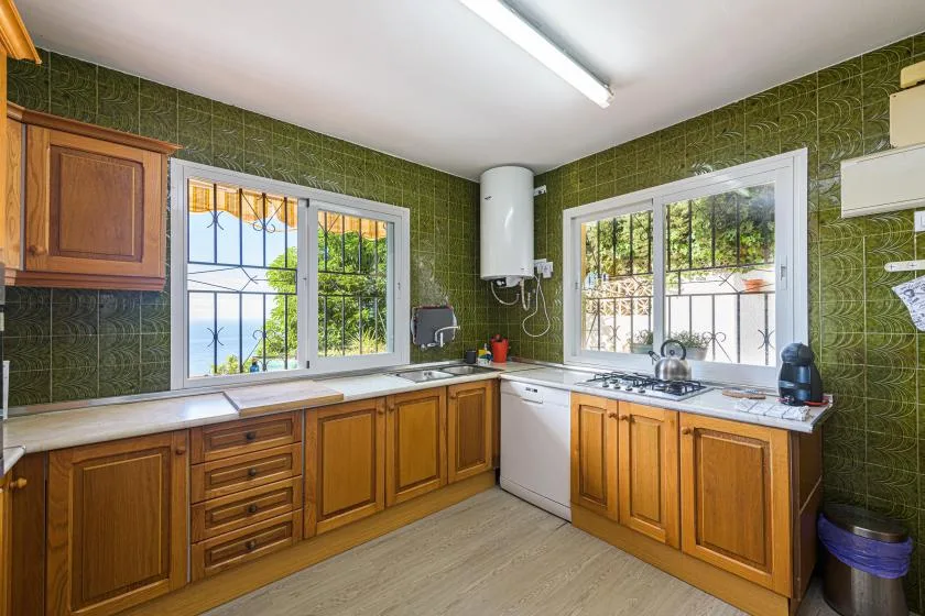 A kitchen with wooden cabinets, green tiled walls, and windows over the counters.