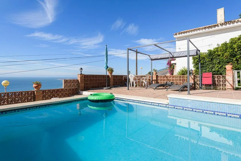 A swimming pool beside a tiled patio, with the sea visible beyond the railing.