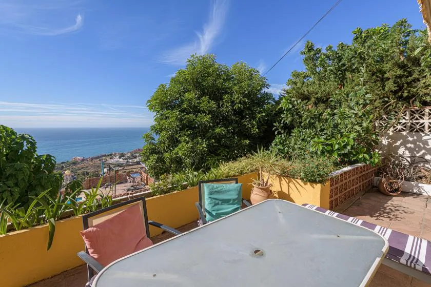 A sunny terrace overlooks the sea, with outdoor seating and a table.