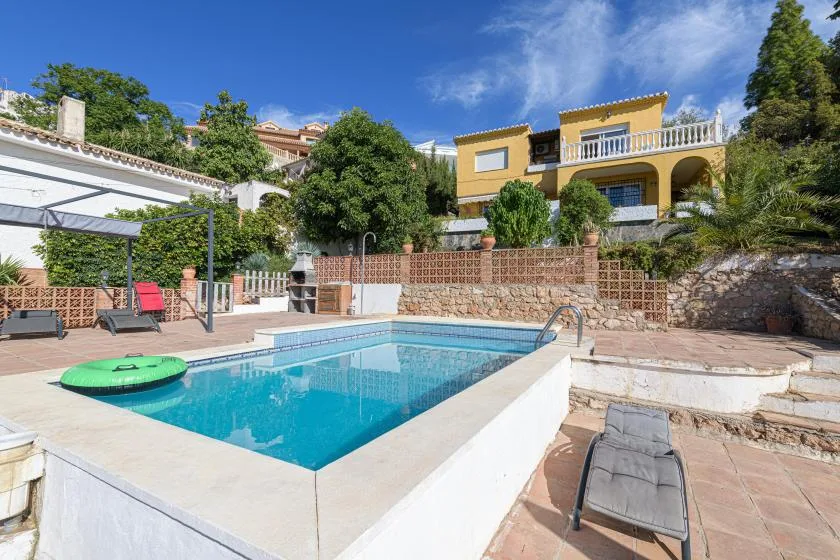 An outdoor swimming pool sits on a sunny patio beside a yellow house.