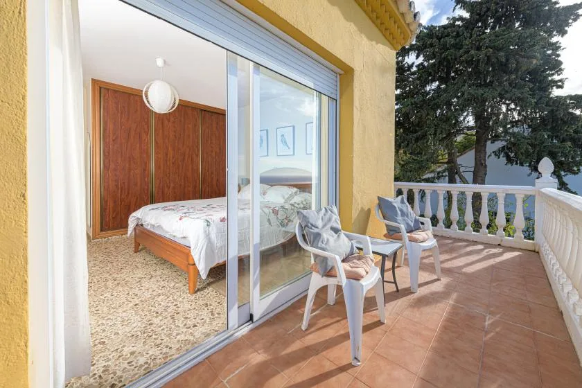 A tiled balcony with outdoor seating overlooks a tree, beside an open bedroom.