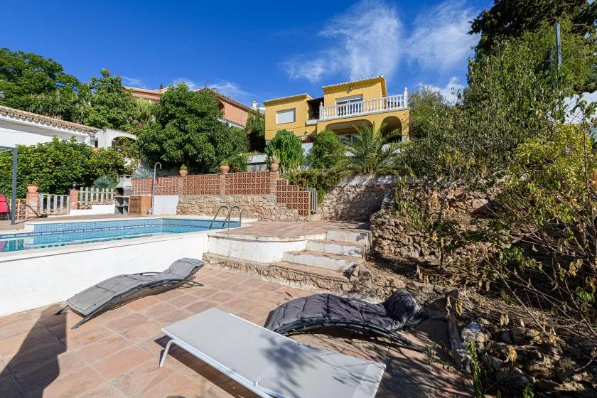 An outdoor pool beside a sunlit tiled patio and landscaped stone wall.