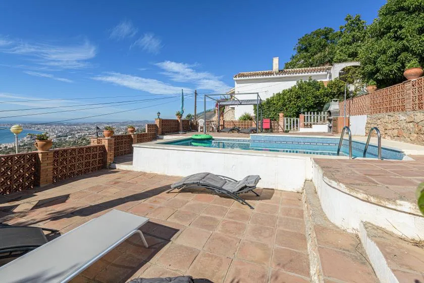 A tiled outdoor terrace surrounds a swimming pool, with lounge seating and a hillside view.