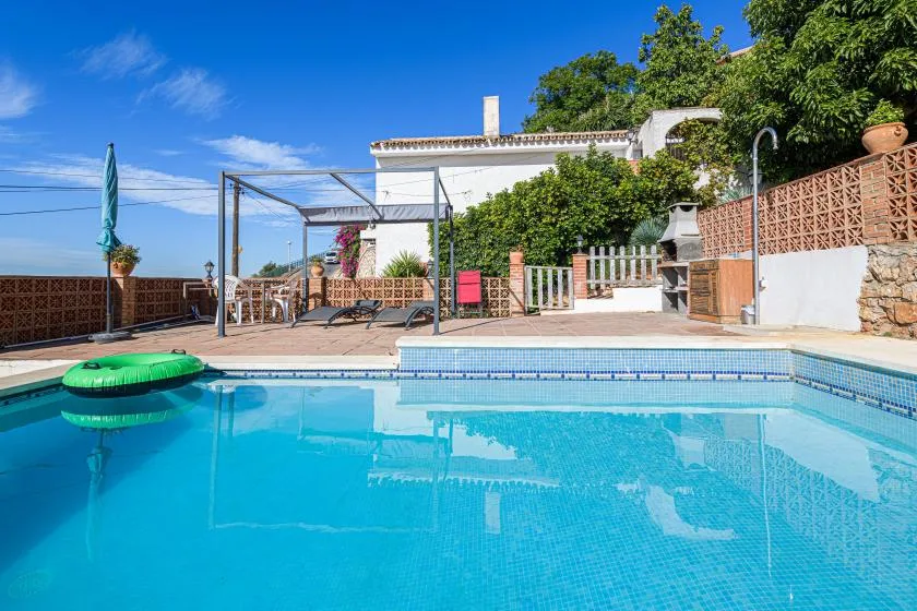 An outdoor swimming pool beside a paved patio and leafy garden.