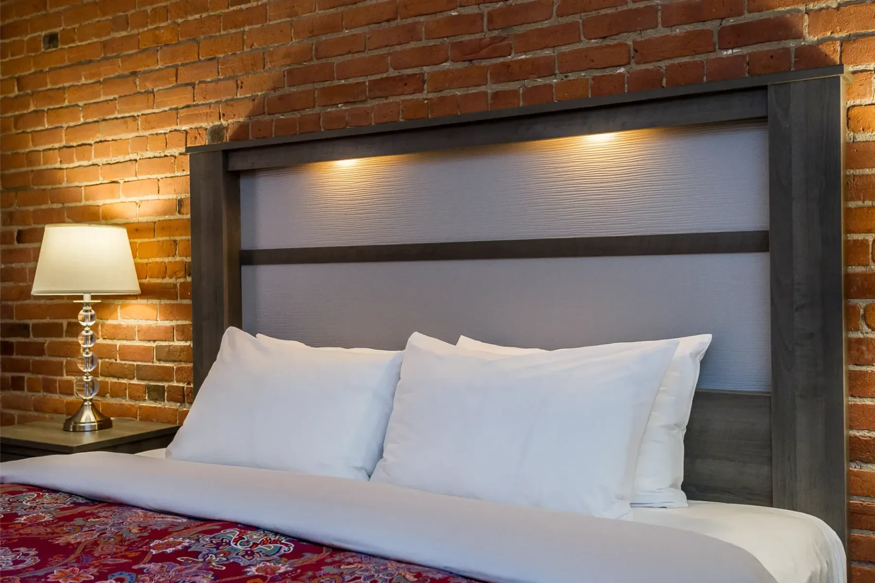 A hotel room bed with an exposed brick wall and lamp.