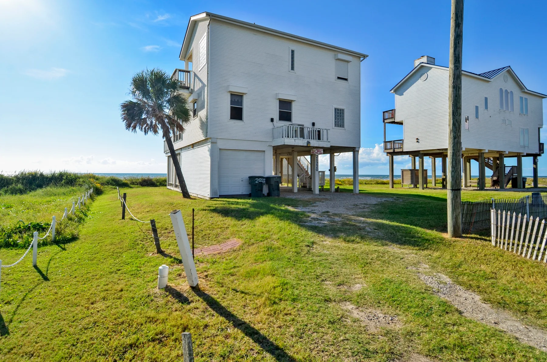 Raised coastal houses stand on supports beside a grassy path.