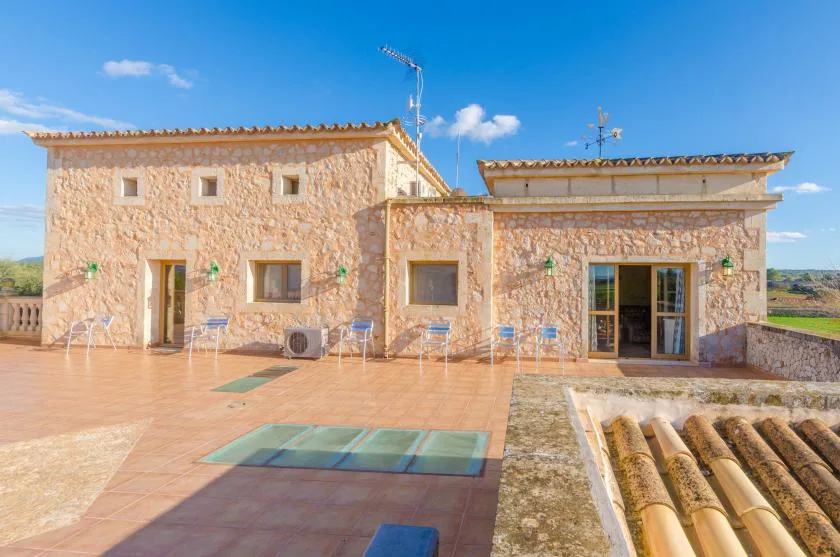 A stone building opens onto a broad tiled terrace beneath a clear blue sky.