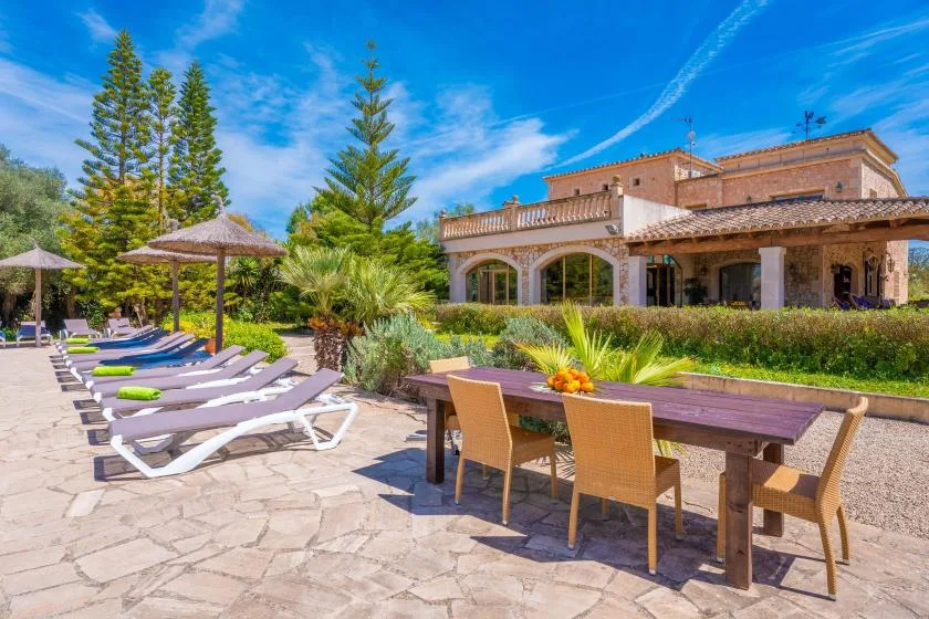 A sunny outdoor patio has lounge seating, a dining table, and a large house behind it.