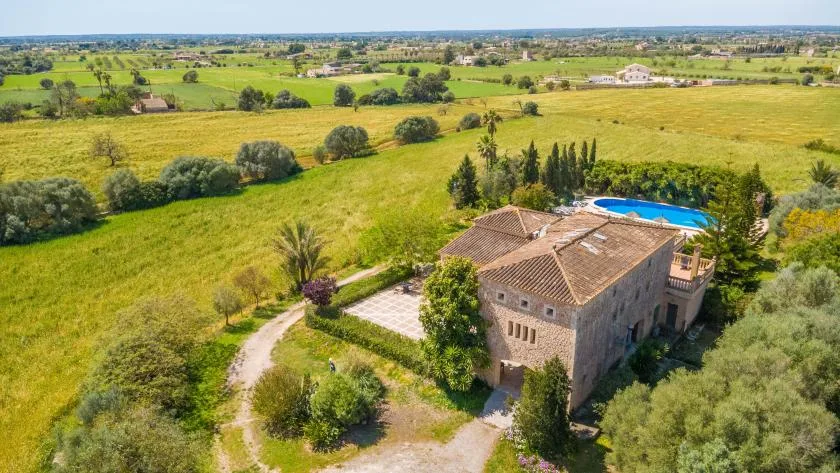 A stone country house with a swimming pool sits amid green and golden fields.