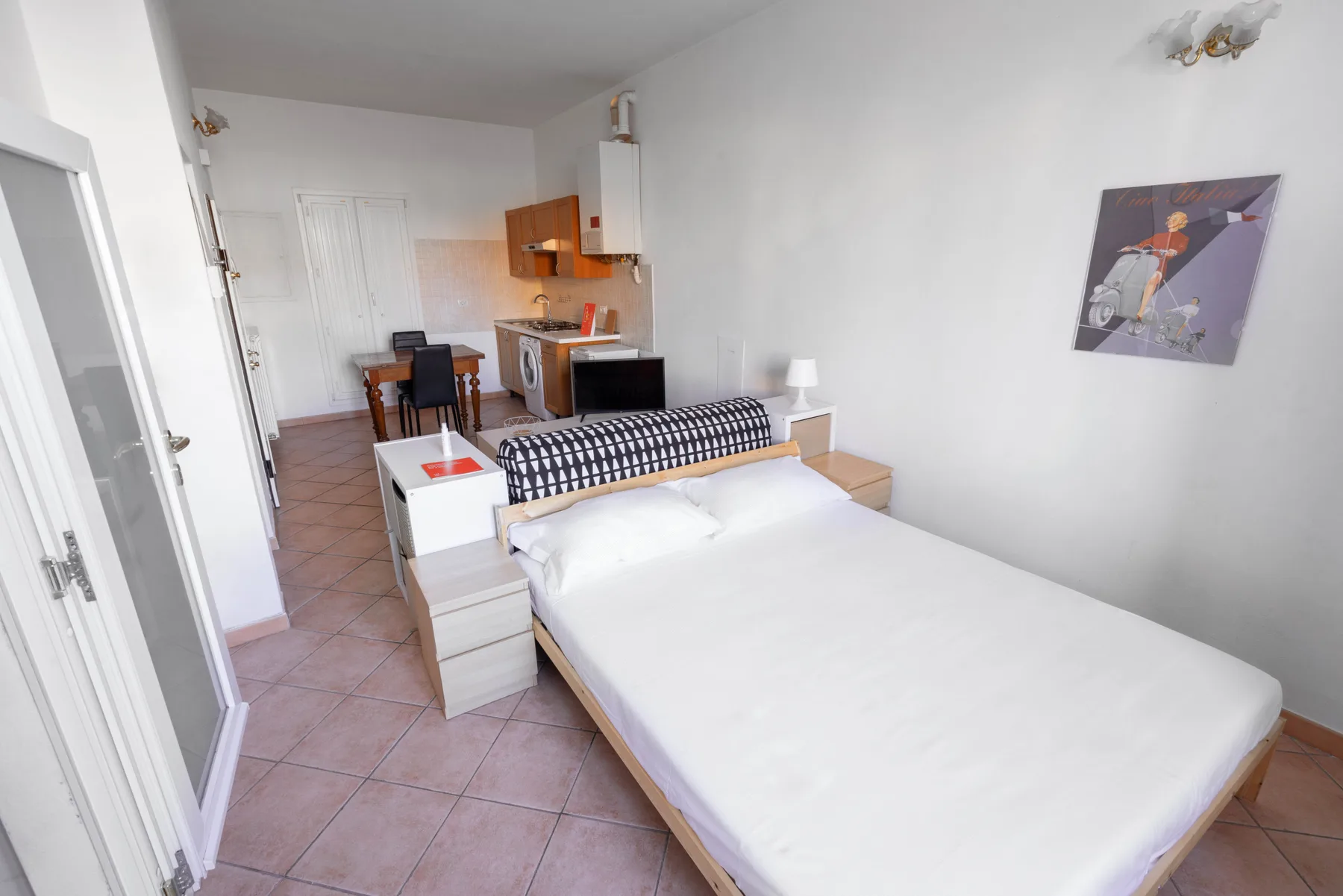 Un dormitorio con una cama, un escritorio y una pequeña cocina al fondo.