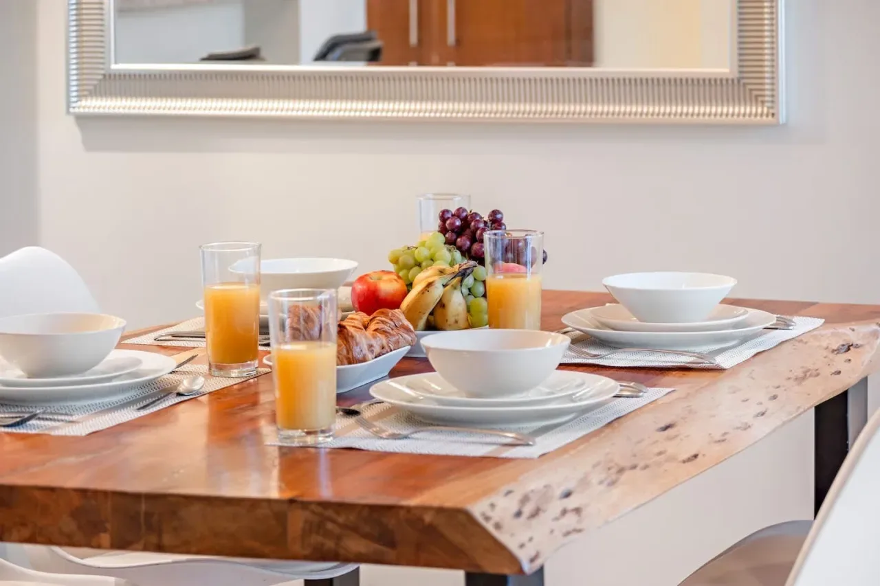 Una mesa de comedor está preparada con platos de desayuno, zumo y fruta.