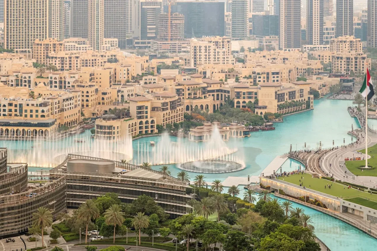 A city skyline overlooks a turquoise waterfront, with fountains and landscaped paths.