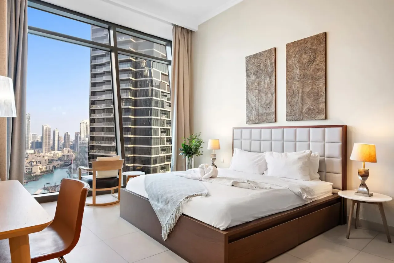 A bright bedroom with a large bed beside floor-to-ceiling windows overlooking city buildings.