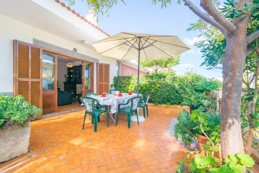 A patio dining area with a table beneath a large umbrella, surrounded by greenery.