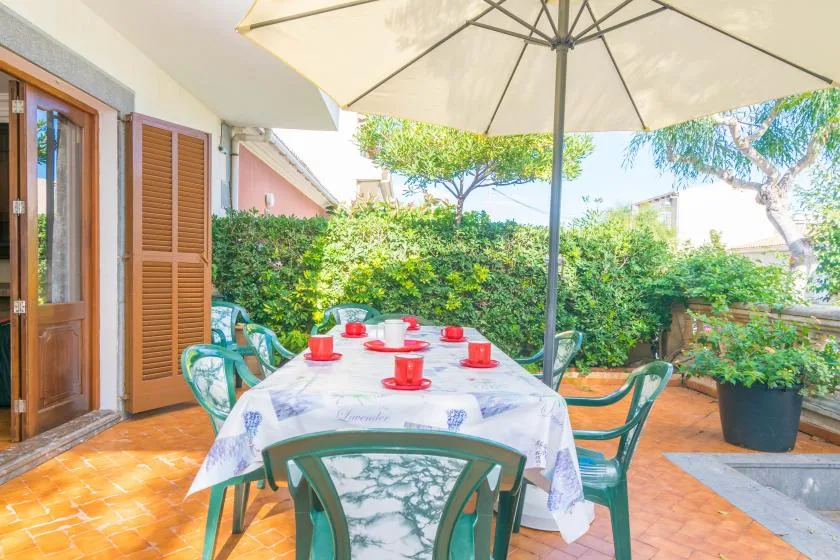 An outdoor dining area with a table beneath a large umbrella, surrounded by greenery.