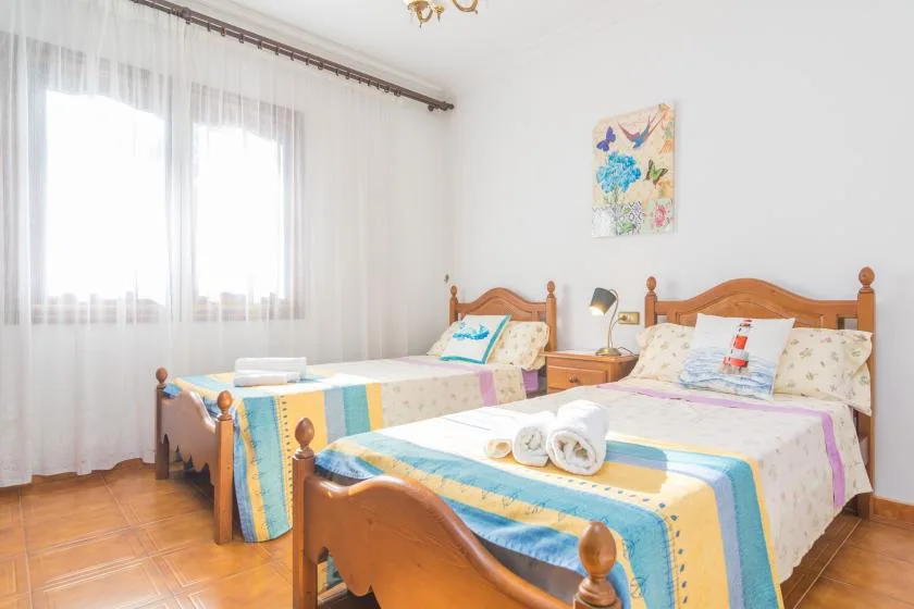 A bright bedroom with two beds dressed in colorful bedding beside a large curtained window.