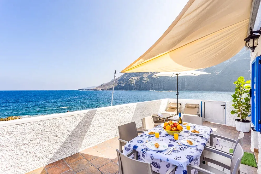 A seaside dining terrace overlooks the water, with a table set beneath a sunshade.