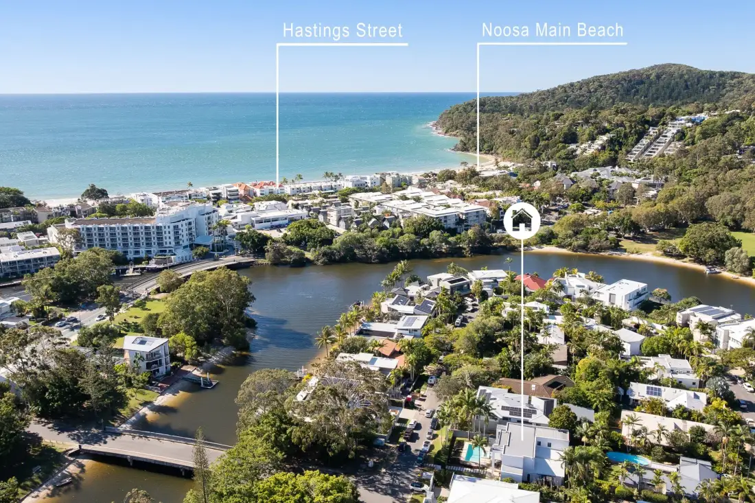 Aerial view of a coastal neighborhood beside a river, with the ocean and wooded hills beyond.