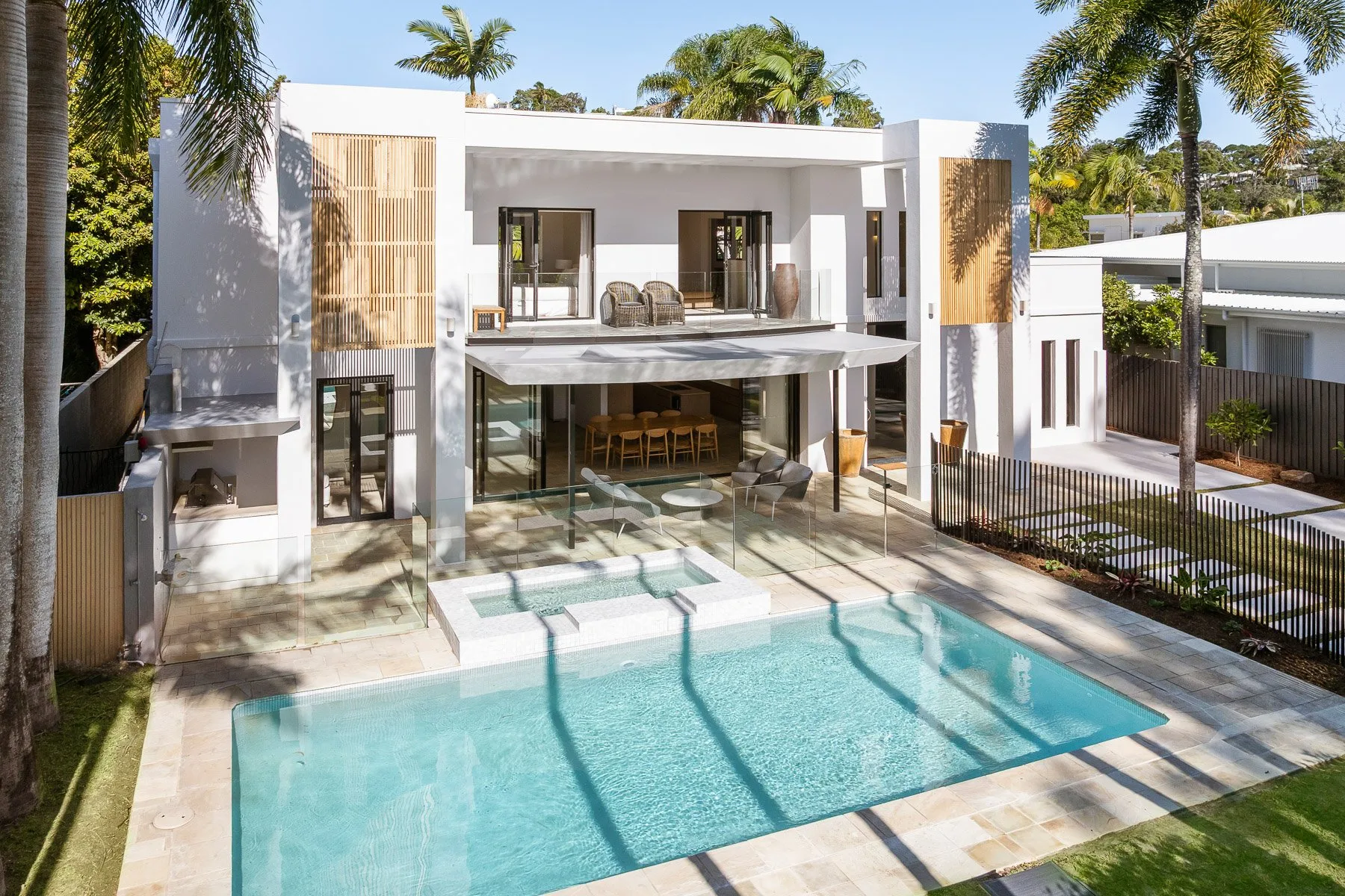 A modern white house overlooks a swimming pool, with palm trees around the property.